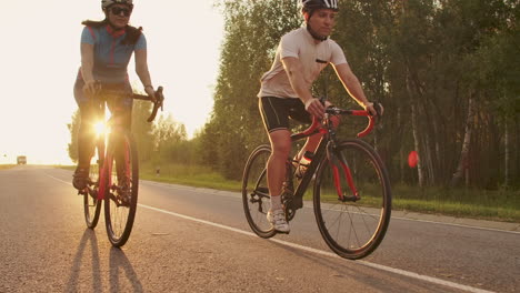 Ein-Mann-Und-Eine-Frau-Fahren-Bei-Sonnenuntergang-In-Zeitlupe-Gemeinsam-Auf-Fahrrädern-Die-Straße-Entlang.-Das-Paar-Ist-Mit-Dem-Fahrrad-Unterwegs-Und-Trägt-Fahrradhelme.